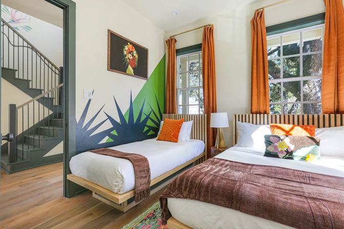 Sunlit vacation-rental bedroom with two platform beds (single and double), striped headboards, orange curtains framing windows with tree views, green geometric wall mural, colorful throw pillows and brown blankets on hardwood floors, staircase visible through open doorway.