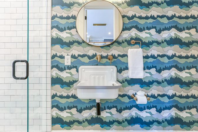 Modern powder room with colorful layered mountain wallpaper, round mirror, wall-mounted white sink, brass fixtures, towel and toilet-paper holders, and glass subway-tile shower door.