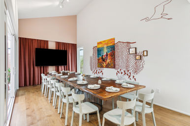 Bright coastal-inspired dining room with a long rustic wooden table set for a crowd, white chairs, hardwood floors, sliding glass doors, maroon curtains behind a wall-mounted TV, and colorful seascape and seagull wall art.