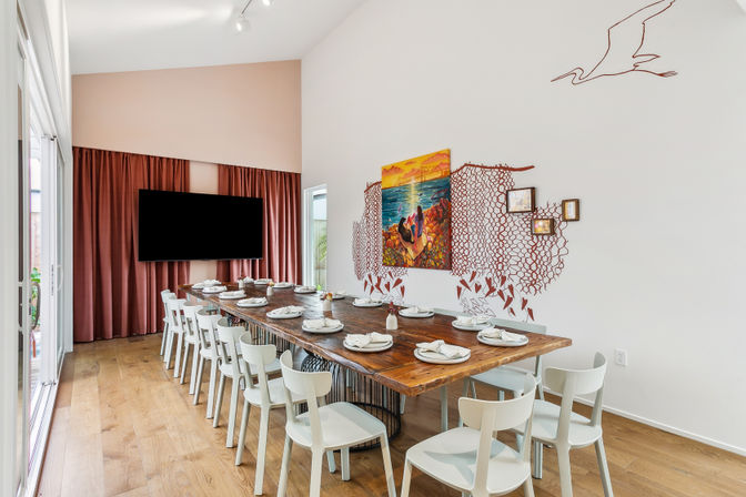 Bright coastal-inspired dining room with a long rustic wooden table set for a crowd, white chairs, hardwood floors, sliding glass doors, maroon curtains behind a wall-mounted TV, and colorful seascape and seagull wall art.