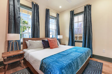 Bright modern bedroom with wood-framed bed, teal blanket and colorful throw pillows, three tall windows with dark blue curtains, mid-century nightstands and lamps.