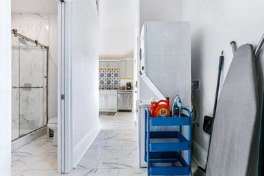 Bright hallway laundry nook with stacked washer-dryer, blue utility cart holding orange laundry detergent and iron, ironing board and broom, marble-tiled bathroom and kitchen with patterned backsplash visible down the hall.
