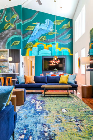 Bright contemporary open-concept living room in an urban loft with high ceilings, large teal mural of a flying blue heron over a city skyline, deep-blue sofa with colorful pillows, vibrant blue-green abstract rug, pendant lights and orange kitchen stools.