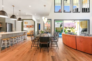Bright open-concept kitchen and dining room with a long island and stools, wooden dining table with black chairs, curved orange sofa, and sliding glass doors to a sunny backyard patio and pool.