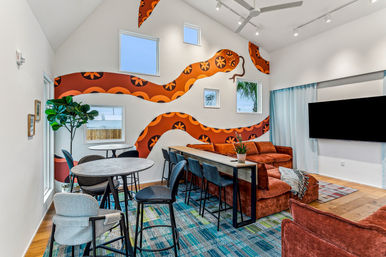 Bright modern communal lounge with bold orange snake-pattern mural, rust velvet sectional, bar-height table and stools, round café tables, flat-screen TV and blue patterned rug