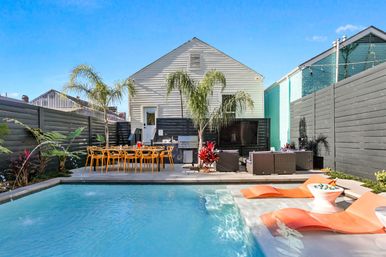 Sunny modern backyard pool patio with shallow tanning ledge and orange loungers, outdoor dining table, wicker lounge area, palm trees and white-sided house with privacy fence