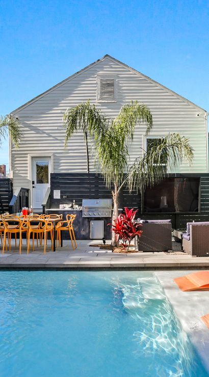 Sunny modern backyard pool patio with shallow tanning ledge and orange loungers, outdoor dining table, wicker lounge area, palm trees and white-sided house with privacy fence