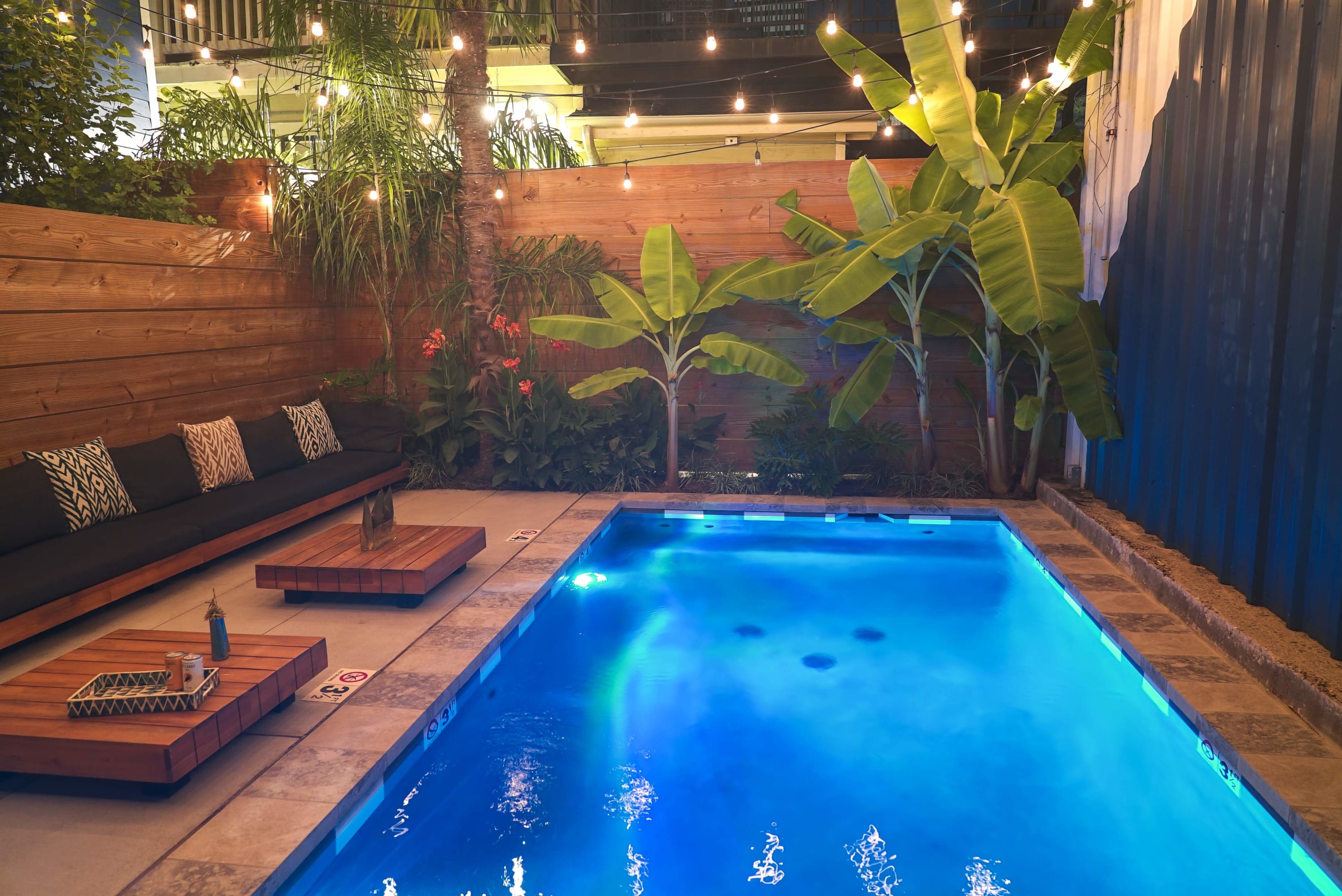 Blue-lit rectangular backyard pool at night with tropical banana plants, string lights overhead, wooden fence and low wooden lounge seating with cushions on a cozy patio