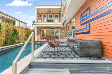 Modern backyard patio with narrow lap pool, bright orange siding with a blue geometric mural, gray modular sofa, woven orange lounge chairs on a patterned outdoor rug, deck steps to the water and a covered second-story balcony with ceiling fan