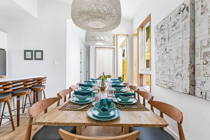 Bright modern dining room with long natural wood table set for eight with turquoise dishes, woven pendant lights, wooden chairs, bar stools and large windows