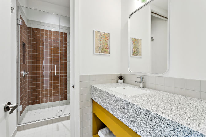 Modern bathroom interior with speckled terrazzo countertop and sink, cheerful yellow vanity, large mirror, glass-enclosed walk-in shower with brown square tiles, and small potted succulent