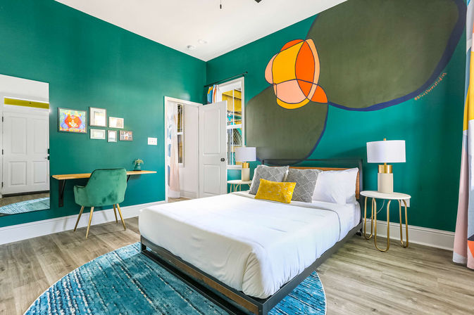 Bright modern bedroom with teal accent wall and colorful abstract mural, white bed with yellow accent pillow, gold-accented nightstands and lamps, green velvet desk chair, round blue rug, and wood-look flooring.