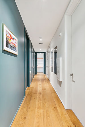Modern urban apartment hallway — long corridor with light oak floors, teal accent wall, recessed ceiling lights, framed artwork and white doors.