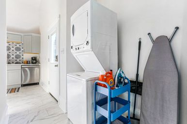 Bright laundry nook with stacked washer-dryer beside a compact kitchen, blue utility cart holding detergent and an iron, gray ironing board and marble-look tile floor.