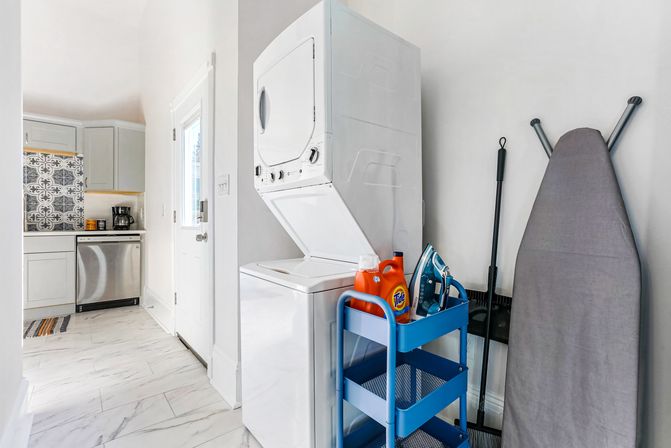 Bright laundry nook with stacked washer-dryer beside a compact kitchen, blue utility cart holding detergent and an iron, gray ironing board and marble-look tile floor.