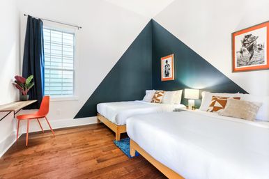 Bright modern guest bedroom with two white beds on hardwood floors, navy triangular accent wall, orange-framed art, bedside lamp, window with shutters, floating desk and orange chair with pops of orange for a playful, clean look