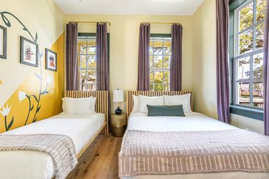 Cheerful sunlit vacation rental bedroom with twin and full beds, yellow floral accent wall, tall windows with purple curtains, wood floors and a bedside lamp.