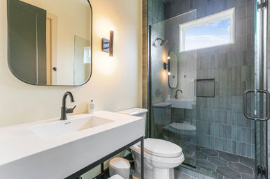 Bright modern bathroom with white vanity, matte black fixtures, rounded rectangular mirror, glass walk-in shower with gray vertical tiles and dark geometric floor tiles