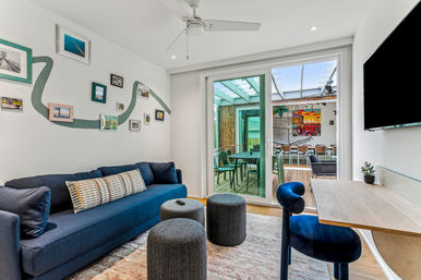 Cozy modern living room with navy sofa, gallery wall and ceiling fan opening via sliding glass door to a covered patio dining area with colorful mural — stylish indoor-outdoor space.