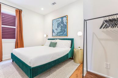 Bright modern bedroom with teal velvet bed and white linens, rust-orange curtains, gold nightstand, framed wall art and open clothing rack