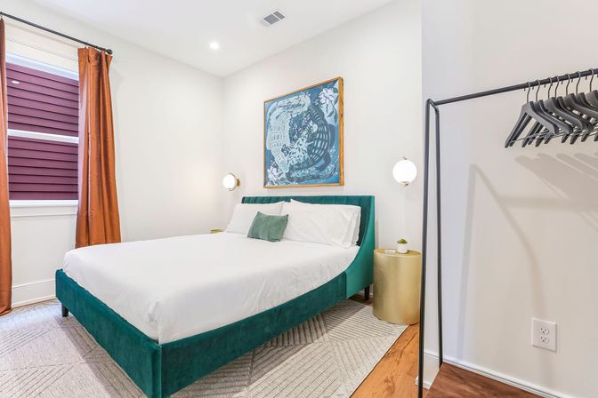 Bright modern bedroom with teal velvet bed and white linens, rust-orange curtains, gold nightstand, framed wall art and open clothing rack