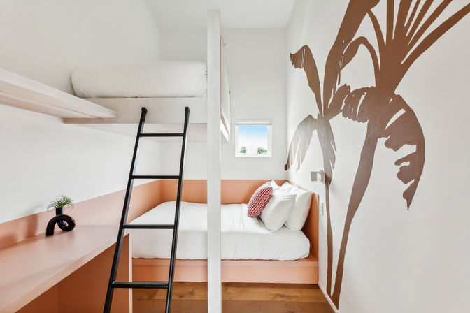 Bright modern bunk bedroom with coral-pink built-in lower bed and white linens, black ladder to upper loft mattress, oversized brown palm tree wall mural, small square window, wood floors and minimalist desk with a plant.