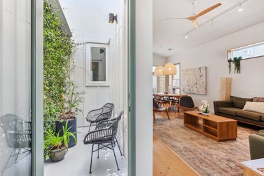 Sunlit narrow courtyard with black wicker chairs and potted plants opening into a modern open-plan living and dining room with wooden furniture, woven pendant lights, ceiling fan and olive-green sofa.