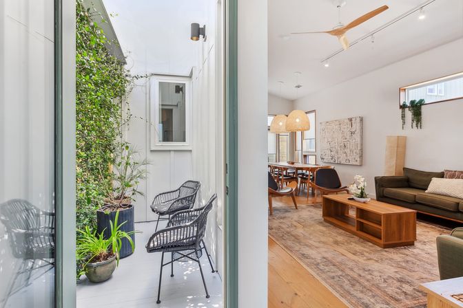 Sunlit narrow courtyard with black wicker chairs and potted plants opening into a modern open-plan living and dining room with wooden furniture, woven pendant lights, ceiling fan and olive-green sofa.