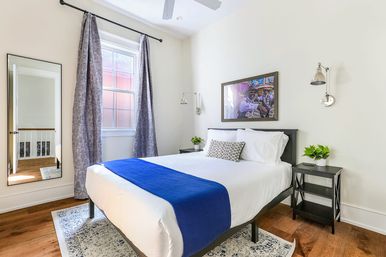 Bright modern bedroom with white walls, queen bed dressed in white linens and blue runner, patterned accent pillow, black nightstands with potted plants, wall-mounted reading lamps, framed art above the headboard, tall window with gray curtains, full-length mirror, ceiling fan and hardwood floors.