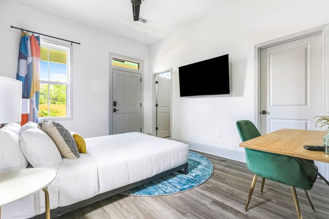 Bright modern bedroom with white walls and wood-look floors, a white-ruled bed with patterned throw pillows, colorful geometric curtains at a sunny window, wall-mounted flat-screen TV, round blue rug, wooden desk and green velvet chair — sleek, airy guest room.