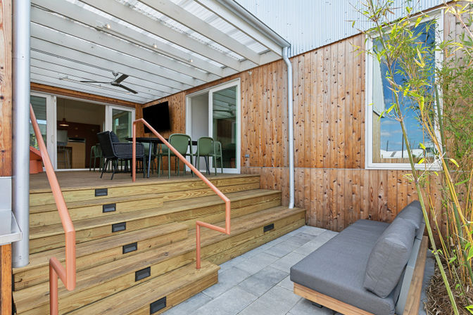 Modern residential patio: covered wooden deck with wide steps and coral metal handrail, outdoor dining set under a translucent pergola with fan, cedar-clad wall, sliding glass doors, gray sofa on stone pavers and bamboo accents.
