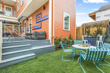 Bright backyard patio with orange siding and blue geometric mural, raised gray deck with lounge chairs, turquoise metal dining chairs around a round table on artificial turf, and a thatched tiki bar.