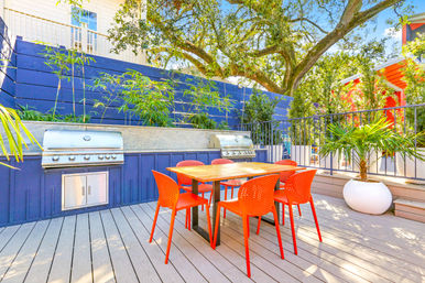 Sunny outdoor deck dining area with six bright orange chairs around a wood table, built-in stainless steel BBQ grills, blue privacy wall and potted palms under a large oak tree.