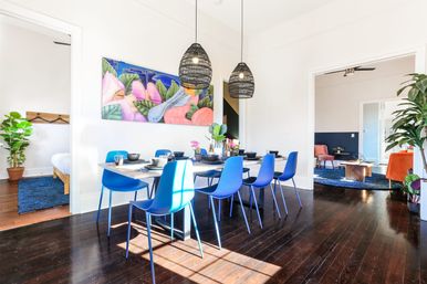 Sunlit modern dining room with blue chairs around a wooden table, woven pendant lights, colorful tropical wall art, indoor plants and dark hardwood floors in an open-plan home.