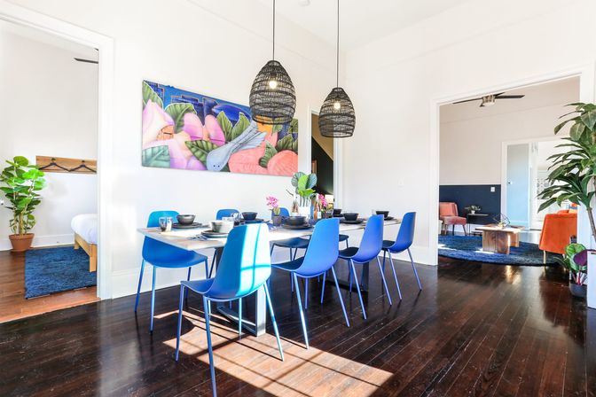 Sunlit modern dining room with blue chairs around a wooden table, woven pendant lights, colorful tropical wall art, indoor plants and dark hardwood floors in an open-plan home.