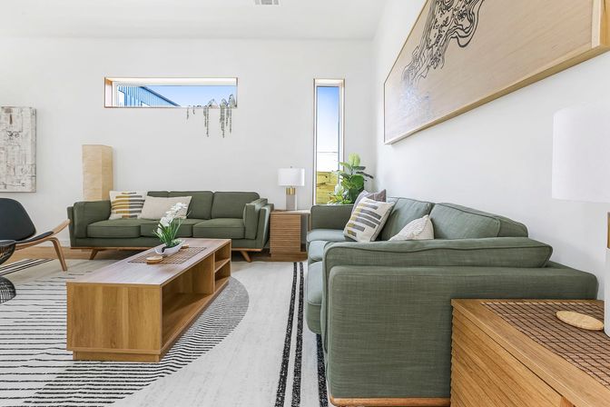 Airy modern living room with olive-green sofas, wooden coffee table and side tables, black-and-white striped rug, potted plant, and abstract wall art