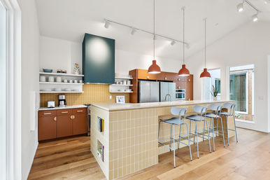 Bright modern open-plan kitchen with a tiled beige breakfast island and built-in shelves, four light-blue metal bar stools, three orange pendant lights, stainless steel fridge and oven, open white shelving with dishes, and warm hardwood floors.