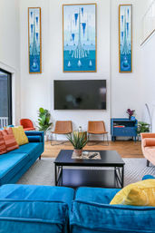 Bright modern living room with high ceiling; wall-mounted TV under three vertical blue abstract paintings, teal velvet sectional with colorful pillows, black coffee table with potted succulent, tan leather chairs, blue side cabinet and indoor plants.