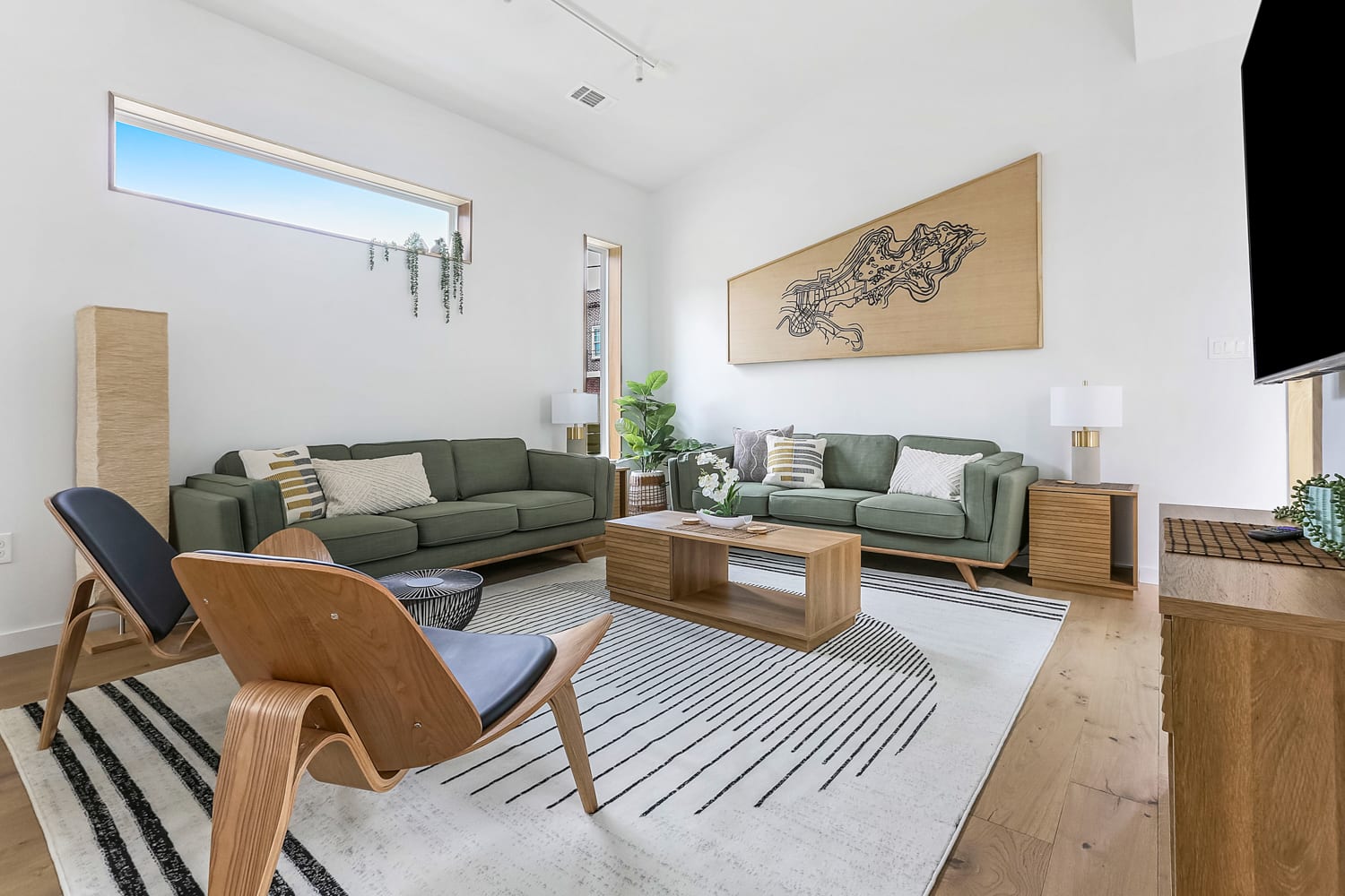 Airy modern living room with mid-century wooden chairs, two green sofas, wooden coffee table, striped rug, indoor plants and abstract wall art beneath a long horizontal window on light hardwood floors.