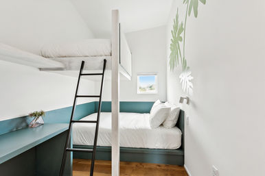 Bright minimalist bedroom with bunk beds: teal built-in lower bed and desk, upper loft mattress reached by black ladder, white linens, small window and green tropical leaf wall decal.