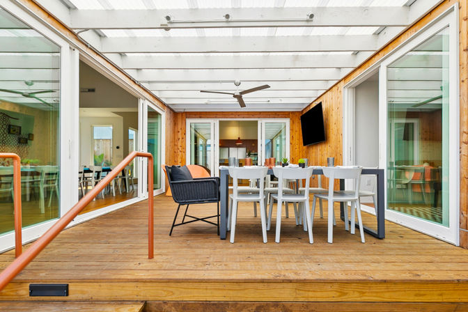 Bright covered wood deck patio with a modern outdoor dining table, white chairs, wicker lounge chair, ceiling fan and wall-mounted TV, framed by sliding glass doors and a translucent pergola roof.