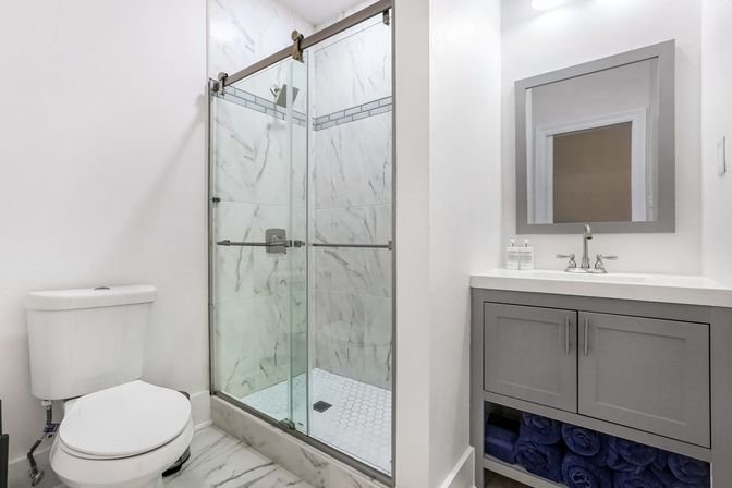 Modern compact bathroom with glass sliding walk-in shower and marble-look tile, white toilet, gray vanity with sink and mirror, rolled navy towels.