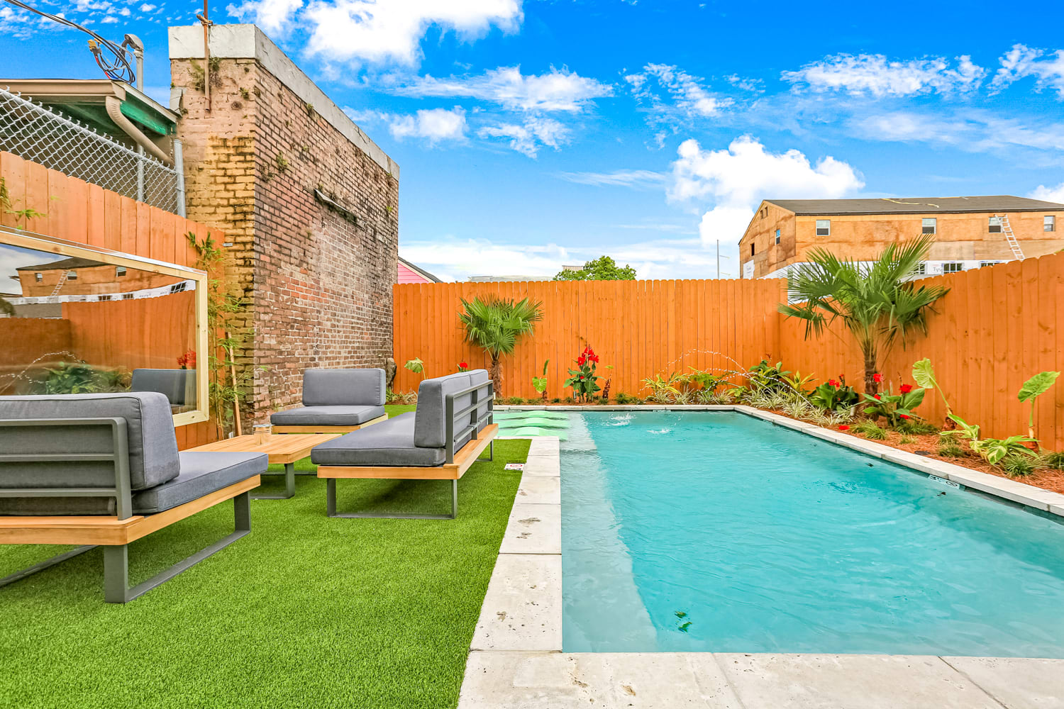 Urban backyard pool with turquoise water, modern gray lounge seating on artificial turf, wooden privacy fence, exposed brick wall and tropical plants under a bright blue sky.