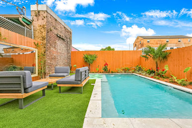 Urban backyard pool with turquoise water, modern gray lounge seating on artificial turf, wooden privacy fence, exposed brick wall and tropical plants under a bright blue sky.