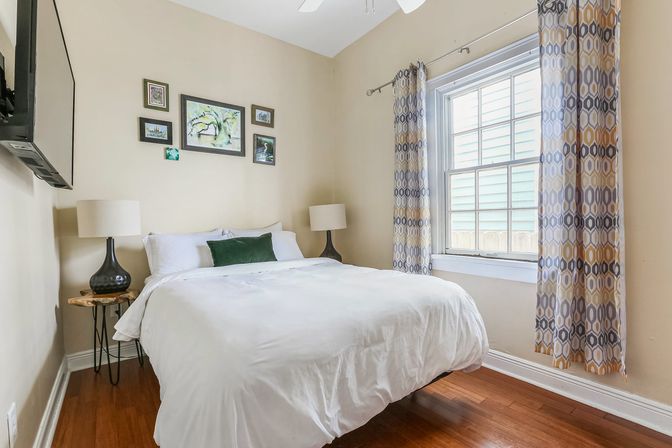 Cozy sunlit suburban bedroom with white bedding and green accent pillow, hardwood floors, patterned curtains, wall art, bedside lamps and wall-mounted TV