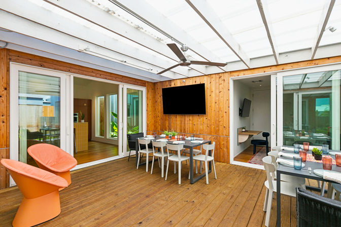 Covered wooden deck patio with sliding glass doors, large ceiling fan and wall-mounted TV; modern outdoor dining table set with plates and glassware, two orange lounge chairs and potted plants.