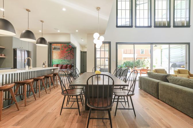 Bright modern open-plan dining and kitchen in a contemporary home — long wooden table with black Windsor-style chairs, kitchen island with wooden barstools and pendant lights, tall windows and sliding glass doors to a sunlit patio, warm hardwood floors.