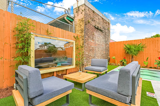 Cozy urban backyard hangout with gray cushioned outdoor seating around a wooden coffee table on artificial turf, exposed brick wall, tall wood fence, tropical plants, and a framed glass panel looking into a small pool under a bright blue sky.