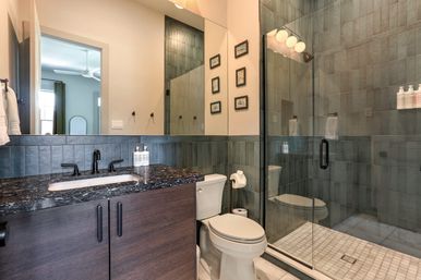 Sleek contemporary bathroom with glass-enclosed tiled shower, dark wood vanity topped with black marble countertop and matte-black faucet, white toilet, large mirror and framed wall art.