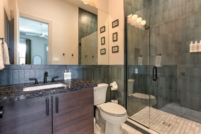 Sleek contemporary bathroom with glass-enclosed tiled shower, dark wood vanity topped with black marble countertop and matte-black faucet, white toilet, large mirror and framed wall art.